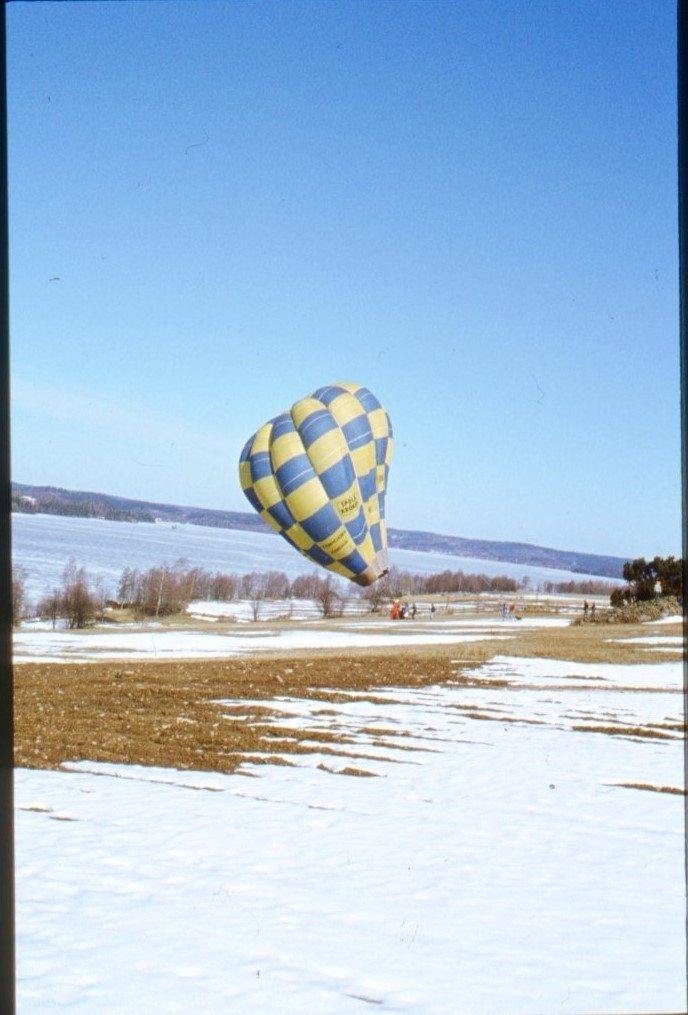 En blågul-rutig ballong med text "Spola kröken" står fylld och klar att lyfta från en delvis snötäckt gräsplan. Isen ligger på sjön i bakgrunden. Ballongen tillhör troligen Tekniska museet.