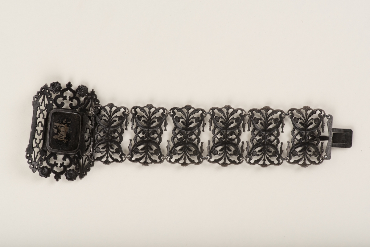 Svart armband tillverkat av järn. Armbandet består av sex ledade delar samt ett spänne. Armbandet är genombrutet och delarna är utformade som motställda liljor med blad. Spännet har en ram i nygotisk stil inom vilken en klassisk figurscen mot en förnicklad bakgrund visas.