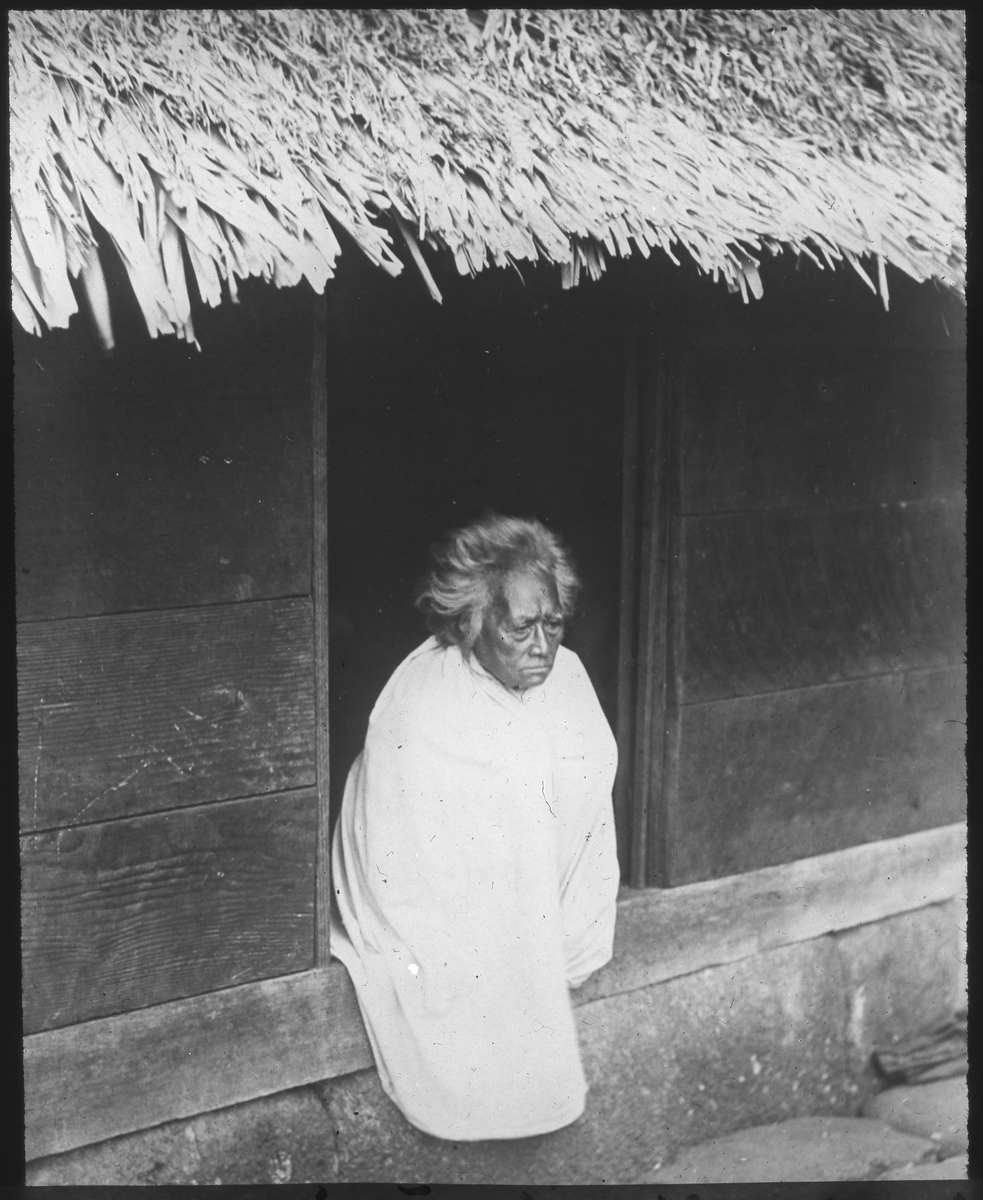 Bilden visar en äldre kvinna på Nuku Hiva klädd i en traditionell dräkt som sitter i ingången till ett trähus. Enlig Etnografiska museets bildinformation är bilden tagen på Fakarava.