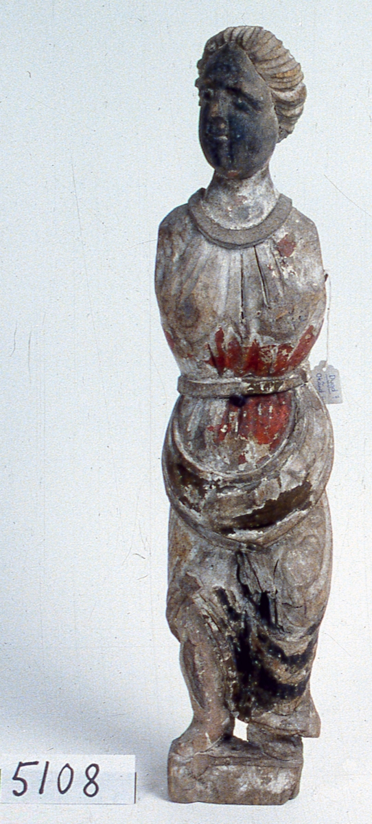 Skulptur föreställer en av dygderna som står frontalt, barfota med lätt utåtböjda knän. Huvudet något högervänt, ansiktet långsmalt, kort bakåtkammat hår. Armar saknas. Klädd i röd blus med rund halsringning. Svart draperad fotsid kjol, hoptagen och sammanhållen med 5-kantigt spänne ovanför höger knä, benet synligt nedanför. Skärp i midjan. Från höfterna och i en båge hänger ett förgyllt draperat tygstycke. Stor kvistspricka vid vänster knä. Vinkelformad urholkning på baksidan. Färgen skadad fläckvis, spår av kredering. Har troligen tillhört altaruppsats eller epitafium. Tillhör samma grupp som 1M16-5107, 96514.