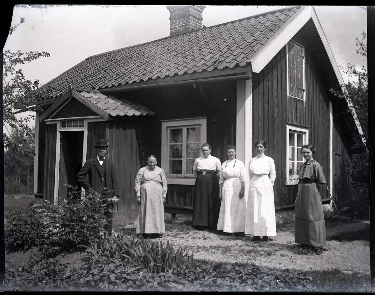 En man i hatt och fem kvinnor stående utanför torp. Trädgårdsland med bl.a. löjtnantshjärta.