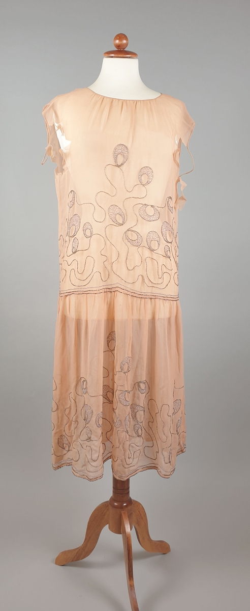 Ferskenfarget, transparent kjole av chiffon med perlebroderier både på liv og på skjørtet. Kjolen er uten ermer og har lav midje. Stoffet er revnet ved begge ermene.