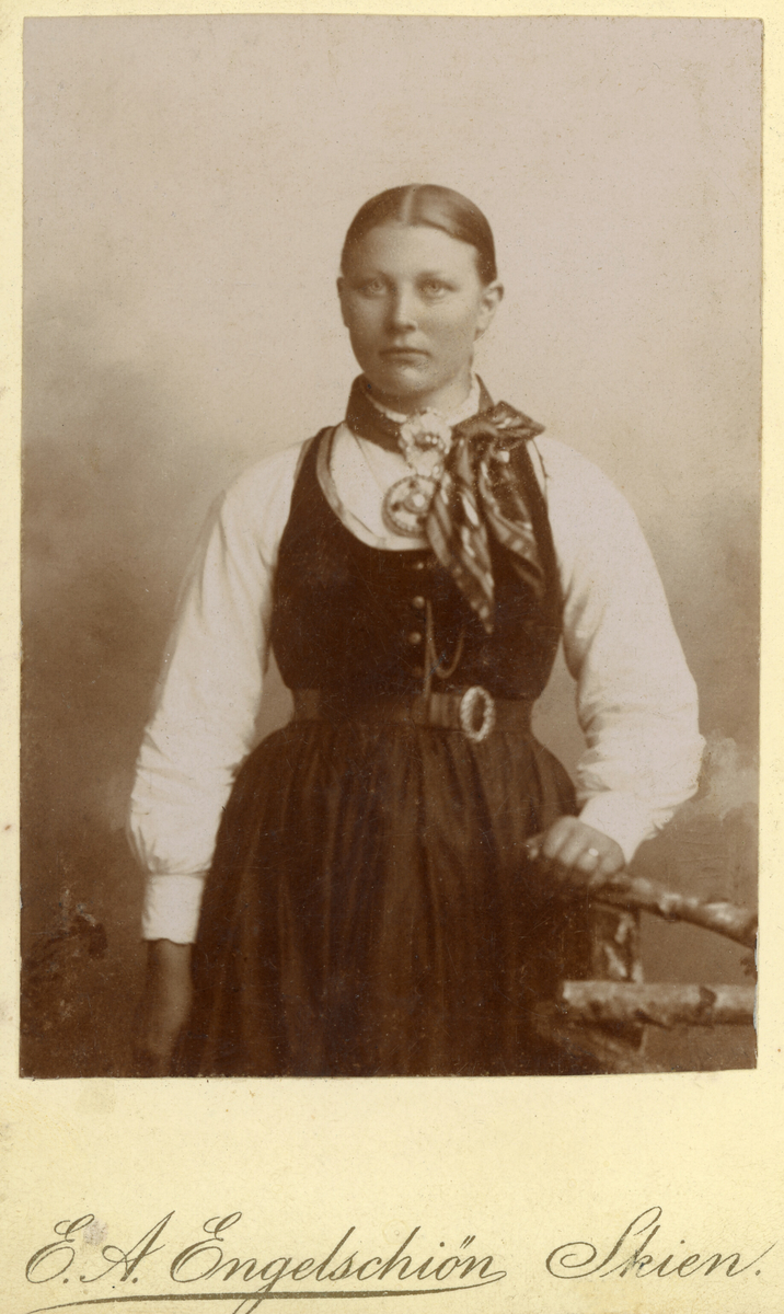 Foto av yngre kvinne i variant av seljorddrakt i atelier.
Kvinna er ukjent.