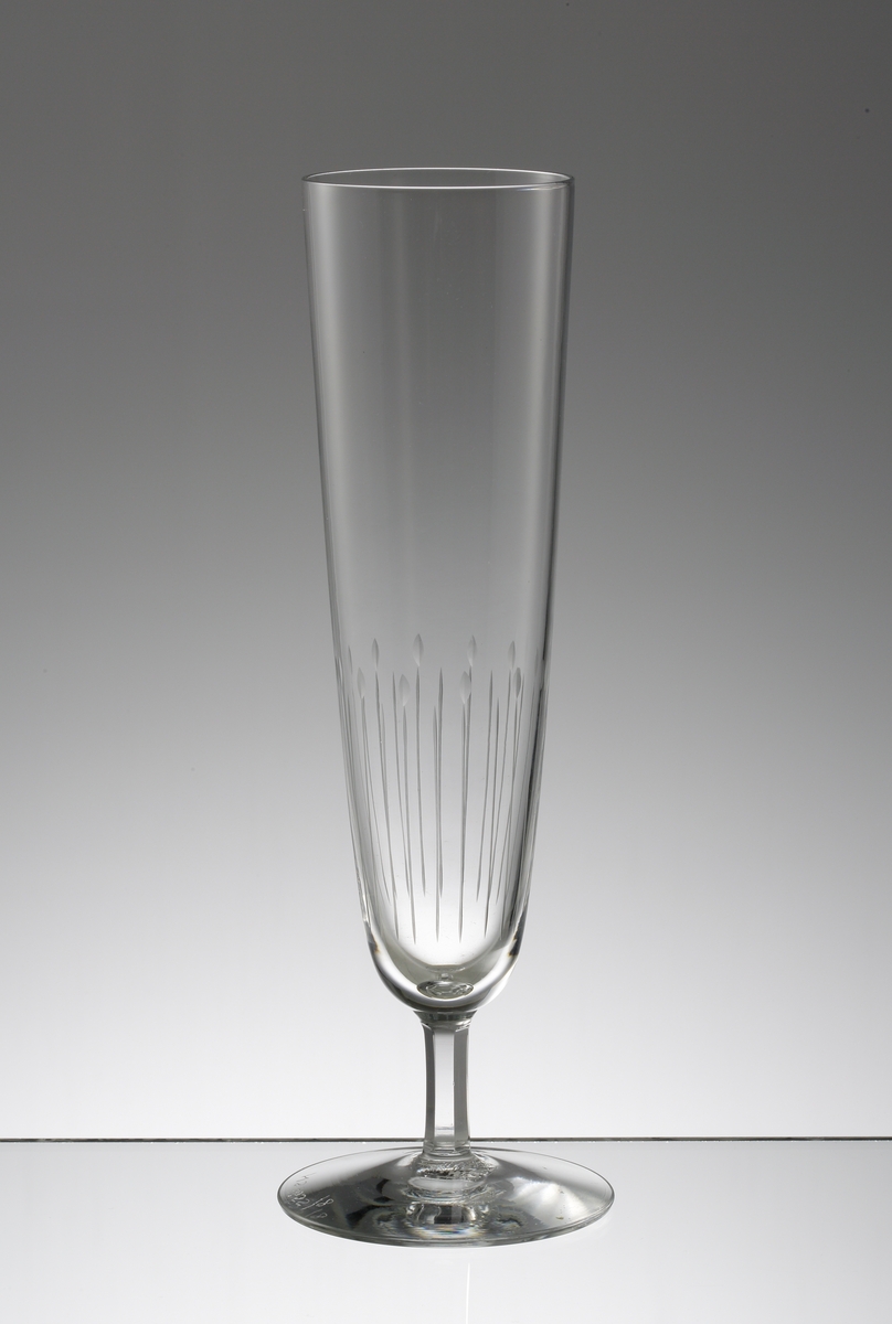 Design: Nils Landberg.
Champagneglas. Hög, svagt konande kupa med lågt fasettslipat ben. Graverad stiliserad dekor på nedre delen av kupan.