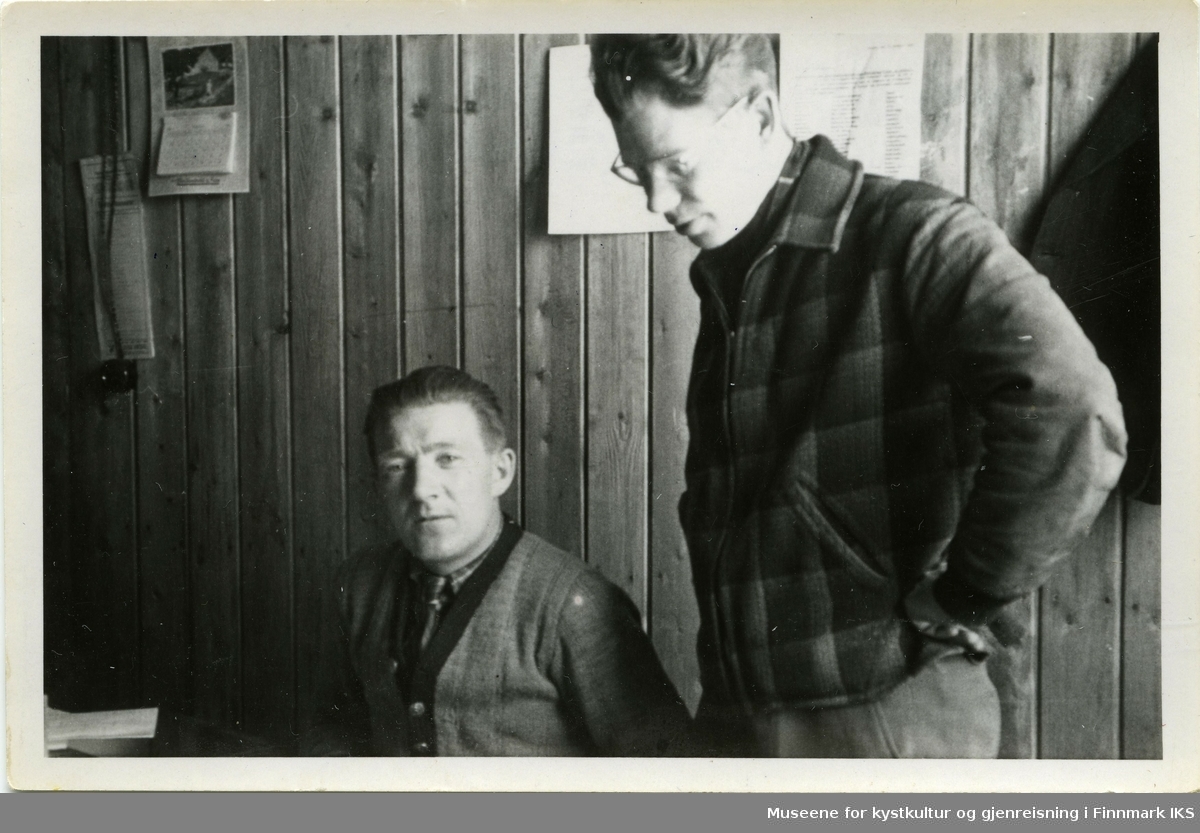 Olaf Berg-Hansen sammen med en annen mann på et brakkekontor i Hammerfest. Mannen til høyre  har på seg briller og ser over skulderen til Berg-Hansen, mens den ser inn i kameraet. Bak på veggen ser man en lysbryter, en kalender og noen dokumenter som er oplslått på veggen.