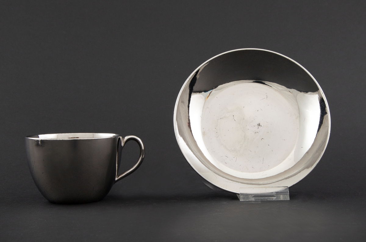 Kopp med skål i keramikk, glasert med sølvfarget luster. Tilnærmet sylinderformet korpus med påsatt hank. Lav sirkulær skål.
