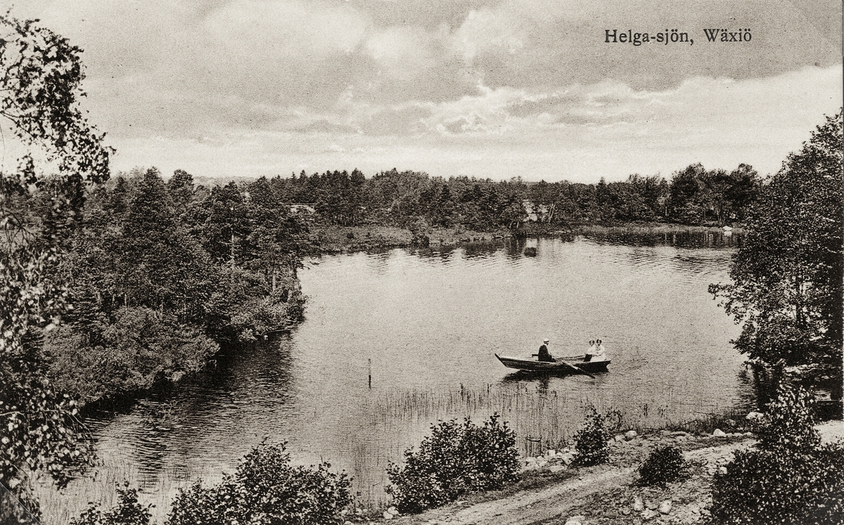 Helgasjön, Växjö, ca 1912. Några personer är ute och ror i en eka. I bakgrunden skymtar några villor bland buskagen.