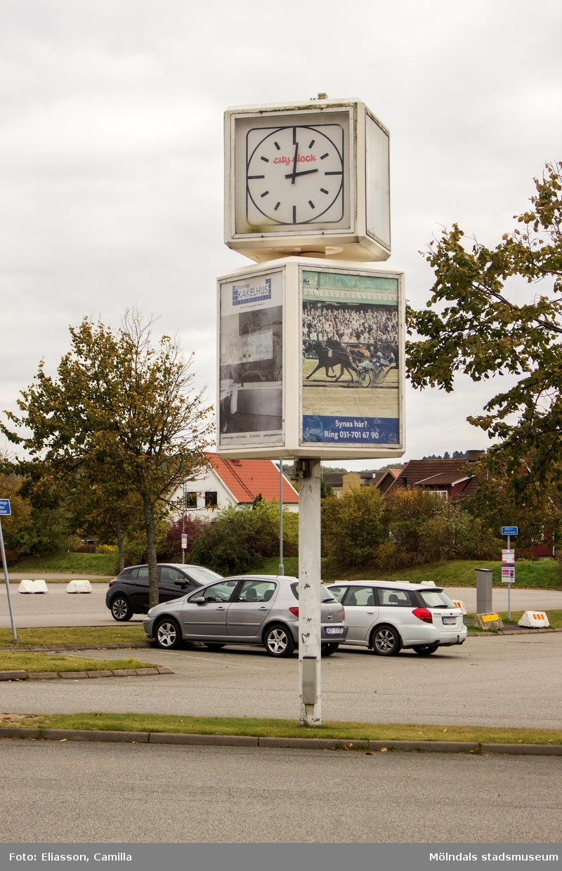 Större klocka på en stolpe vid parkeringsplats tillhörande Åbytravet i Åby, Mölndal, den 11 oktober 2016. Stolpen har även plats för reklam.