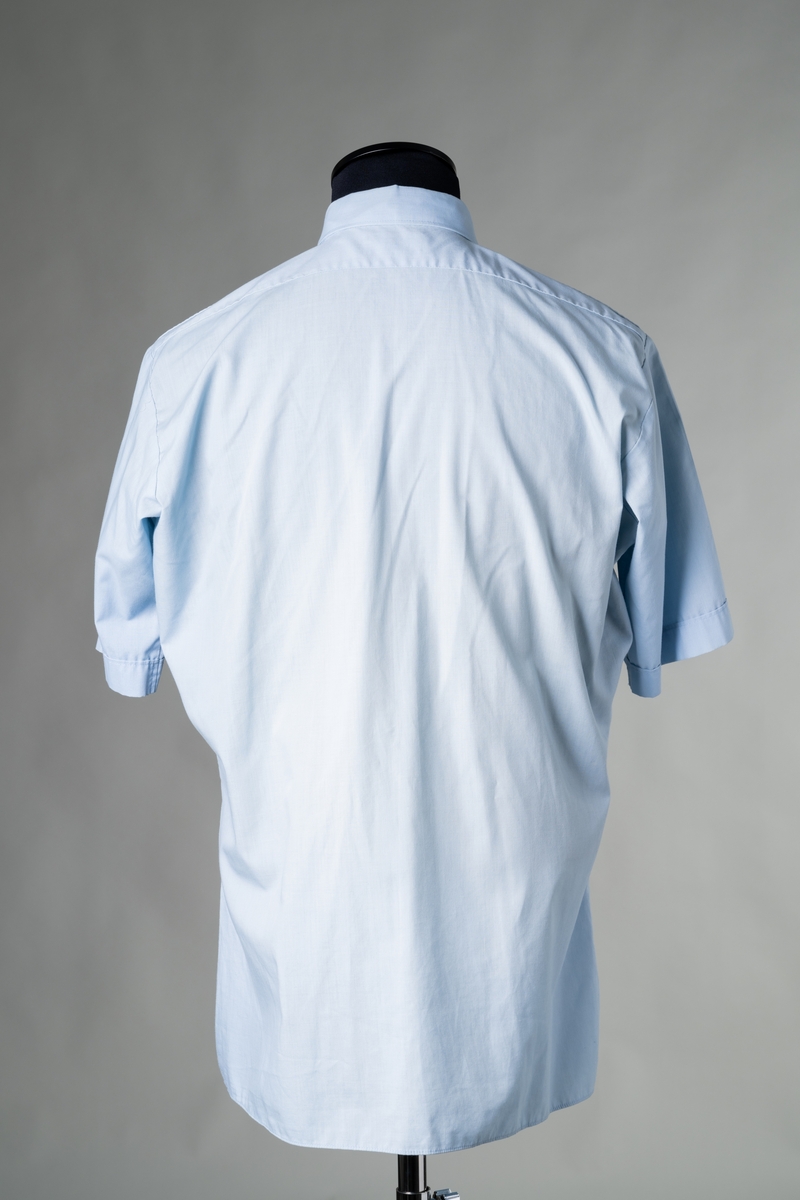 Skjorta till stationsuniform, modell sommar, för personal vid Brandförsvaret, Jönköpings kommun. Kortärmad skjorta av ljusblått tyg av polyester och bomull, insvängd midja, fast krage, ok av dubbelt tyg på ryggen. Knäppning fram med sju vita plastknappar, axelklaffar med en vit plastknapp, och två bröstfickor med lock och vit plastknapp. Tygetikett i nacken: "Bagutta". Storlek: C-bål, L, 42-42.

Samhör med: JM 56718:1-6.

Se vidare Historik.