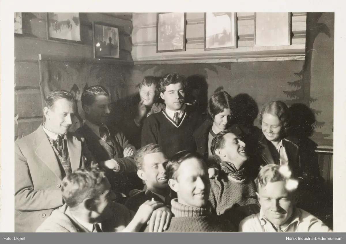 Sollitur, bededagene 1931. Unge mennesker innendørs i lystig lag. Både kvinner og menn sitter trangt sammen i hjørne. Bilder henger langs vegg. Noen røyker en pipe, andre smiler.