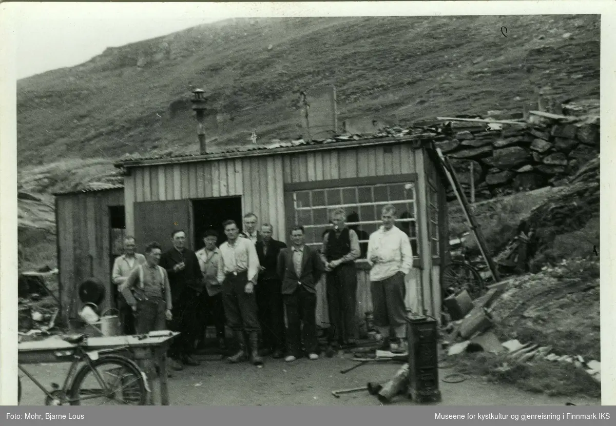 Gruppebilde av ti menn foran en brakke i Hammerfest. Mannen med hvit skjorte som står i midten, er Hans Heggelund. Til venstre står et bord og en sykkel. Til høyre av brakken ser man en mur og i bakgrunnen ligger Salenfjellet.