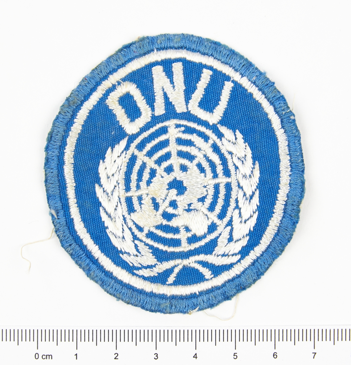 Förbandstecken. Textilt märke, ljusblått, med broderat vitt FN-emblem samt texten ONU.