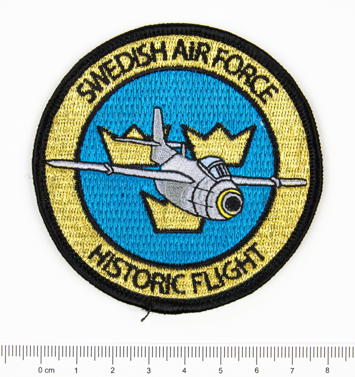Sytt emblem, runt tygmärke med texten Swedish air force historic flight och en "flygande tunna" i mitten framför tre kronor.