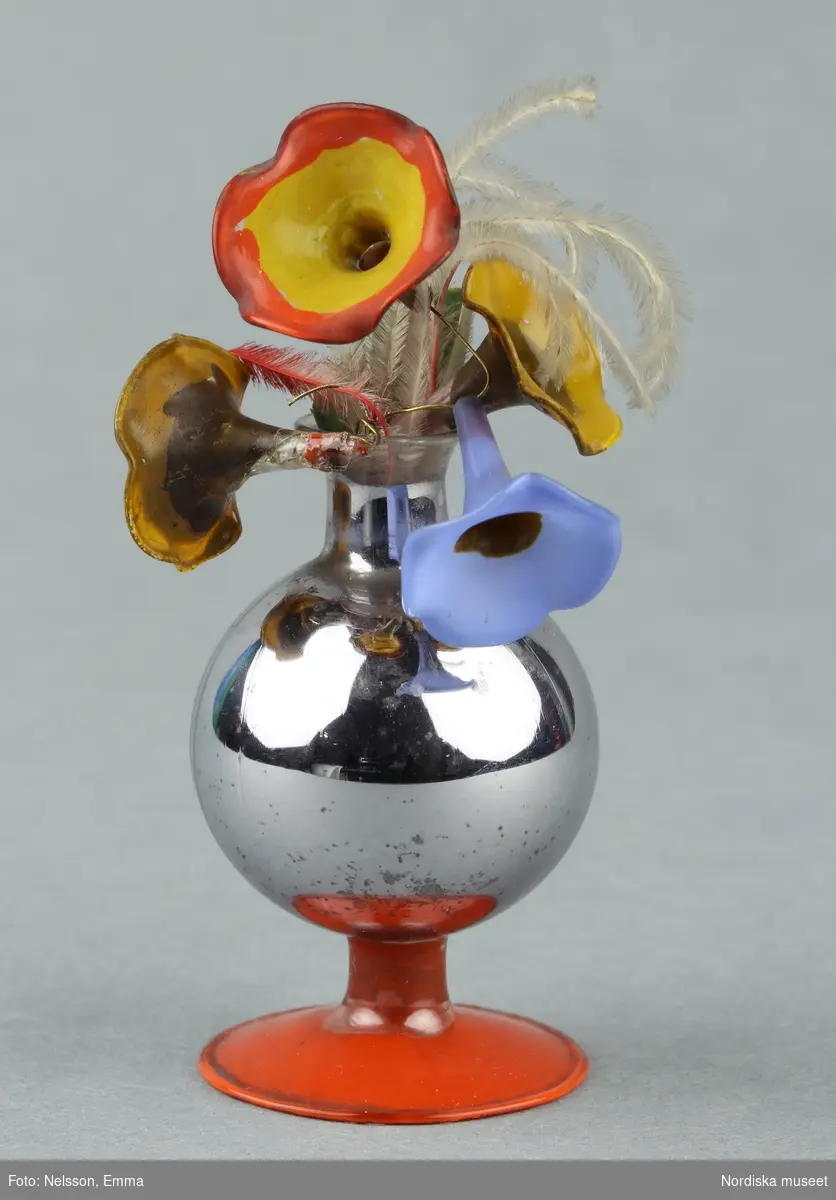 Huvudliggaren:
" Vaser, ett par, leksaker av glas folierat, m. konstgjorda blommor, delvis av glas."Inventering Sesam 1996-1999
H 7,5, Diam 3 cm
Två vaser med blommor. a) vas av kvicksilverglas dvs. folierat glas, rödmålad fot, liljeliknande blommor av flerfärgat glas, dekorerat med tunna fjädrar. b) vas av kvicksilverglas, rödmålad fot, liljeliknande blommor av glas, blommor av textil. Vas b) har sönderslagen fot.  Hör till dockskåp 165280.
Bilaga
Helena Carlsson 1996