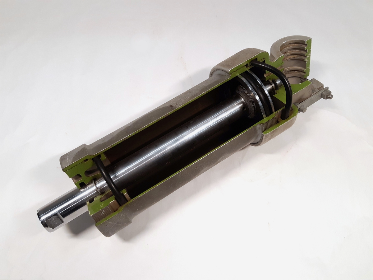 Hydraulcylinder i metall. Uppskuren modell med gröna färgfält. Vid sidan en gjuten märkning: "SM 1031872".