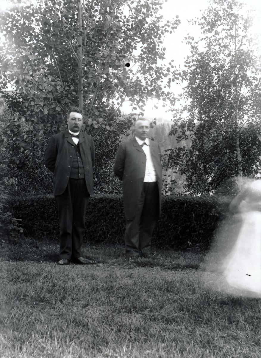 Bestyrer Wilhelm Rydgren, til høyre, sannsynligvis med sin yngre bror Thorvald Rydgren i hagen utenfor bestyrerboligen ved Kistefos Træsliberi.  De står foran klippet hekk iført dressantrekk med skjorte, vest, bonjour og lange bukser. Liten jente i bevegelse i lys kjole i forgrunnen.