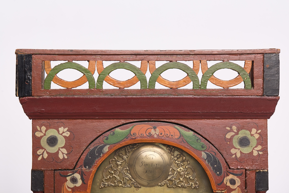 Mørk brunrød, dekorert med påmalte blomster på døra og på toppen. 
(a) kasse
(b) topp/ hode
(c) urverk  - urskive av metall med romerske tall. 
(d) pendel 
(e og f) lodd
(g) sveiv
(h) øvre pyntelist
(i) sokkel