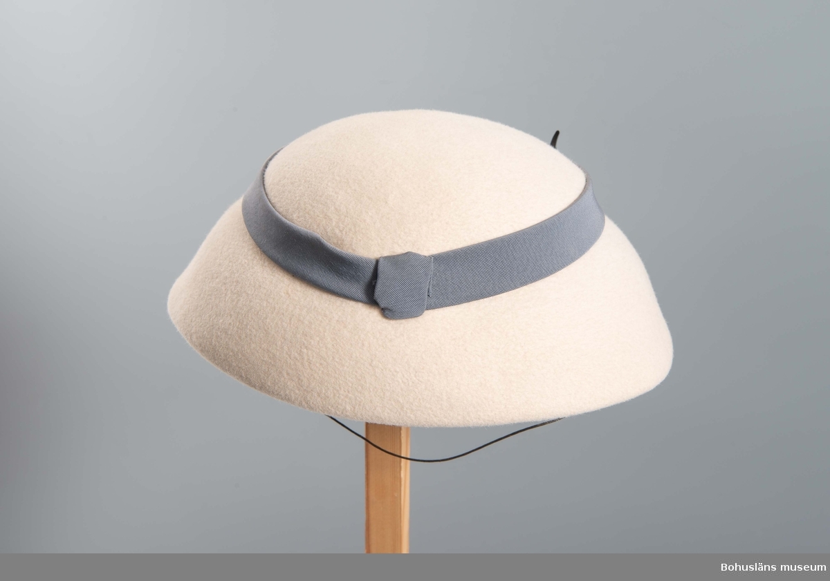 Närmast halvsfärisk hatt i beige filt med svagt antydd kulle kring vilken fästs blågrått ripsband.