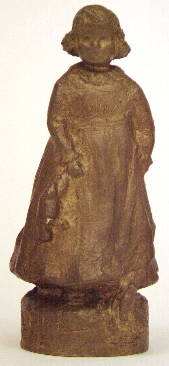 Skulptur eller statyett av brons, "En solstråle" av Ida Matton. Signerad Ida Matton, Paris. Liten flicka med lockigt hår, fotsid klänning med rosett bak. Håller en nallebjörn i höger hand. En docka ligger framför vänstra foten.