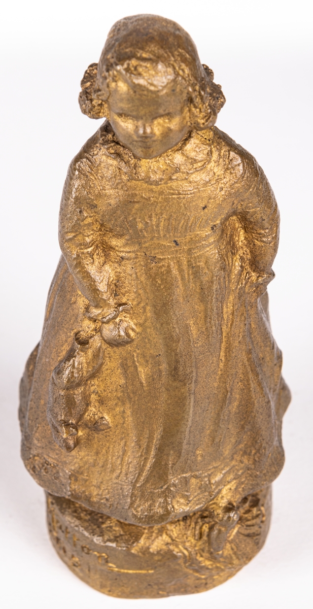 Skulptur eller statyett av brons, "En solstråle" av Ida Matton. Signerad Ida Matton, Paris. Liten flicka med lockigt hår, fotsid klänning med rosett bak. Håller en nallebjörn i höger hand. En docka ligger framför vänstra foten.