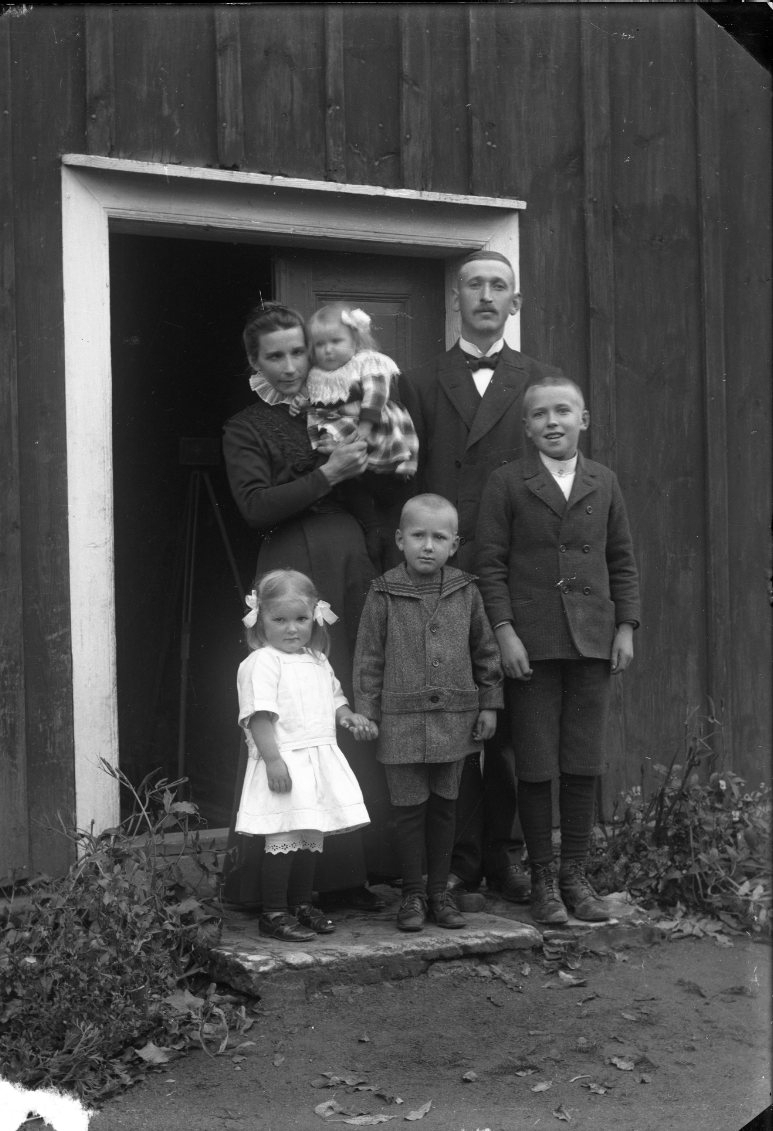 En familj står vid dörren till en stuga. Kvinnan håller ett litet barn i famnen. Till höger om henne står en man och framför dem en liten flicka och två något äldre pojkar. Sannolikt Axel Emil Johansson med familj i Björkeberg, Ölmstad socken.