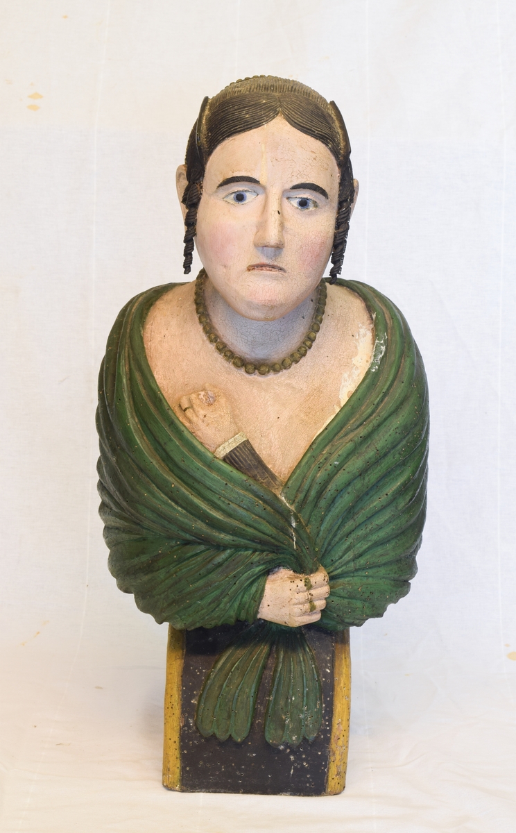 Portrettbyste av kvinnehode. Skulle brukes som gallionsfigurer men kan og ha vært brukt som dekorasjon i et hus. Sortmalt hår med hengekrøller ved ørene og grønt sjal.