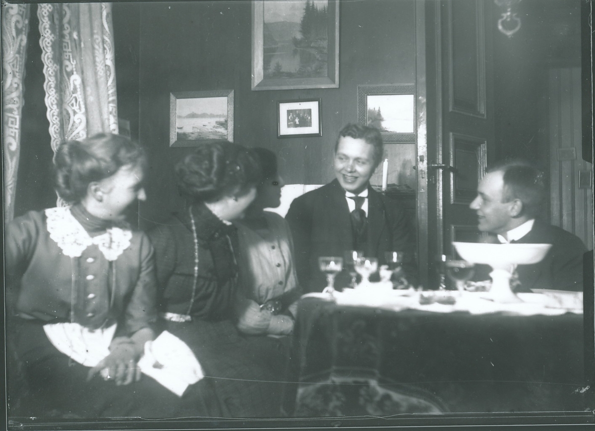 Tre unge kvinner og to unge menn i stueinteriør. Harald Rydgren er nr. 4 fra venstre. De unge menneskene sitter rundt stuebord med fløyelsduk, glass og stettefat. Malerier og fotografier på veggen bak, og blondegardin i vinduet.