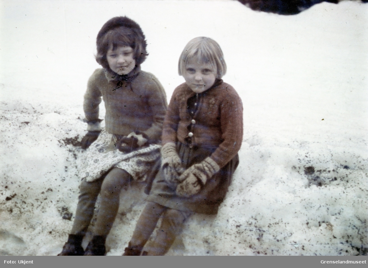 To unge jenter sitter på en snøkant i et snødekt landskap. 