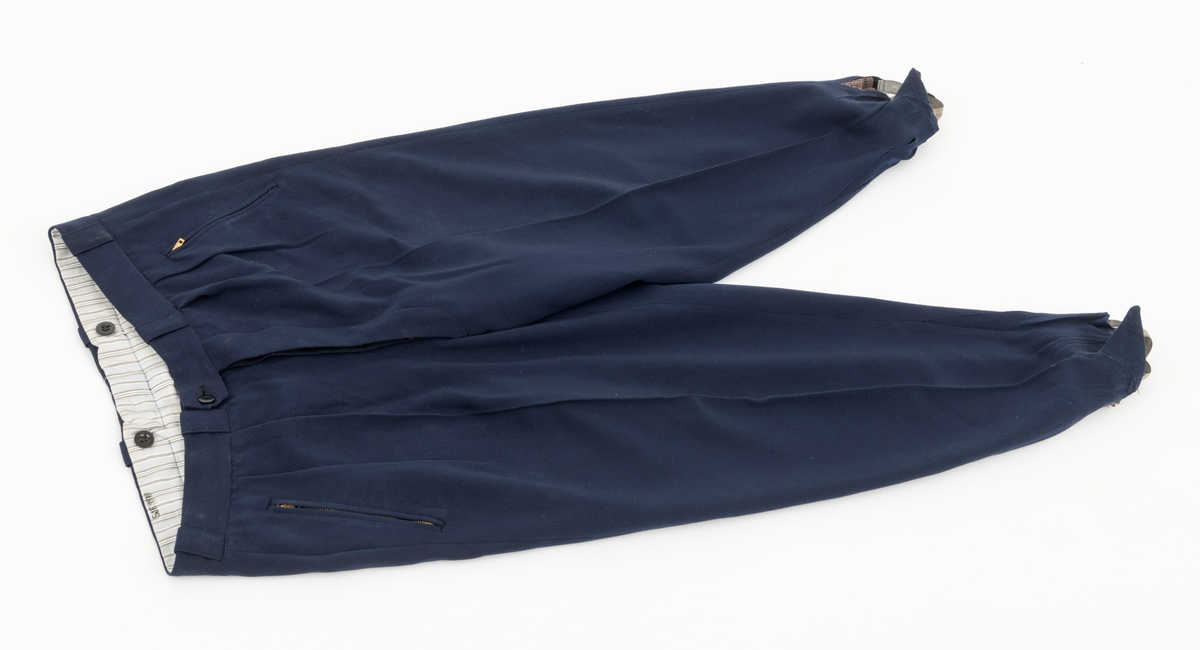 Strekkbukse til skidress i mørkeblå gabardin. Buksa har strikk for å ha under foten. To lommer ved hoftehøyde som lukkes igjen med glidelås. To lommer bak som lukkes igjen med knapper. Buksesmekken lukkes igjen med knapper. Bukselinningen er foret med bomullsstoff. Her er det to knapper foran og to bak til bruk av bukseseler. Buksa har også beltestropper.