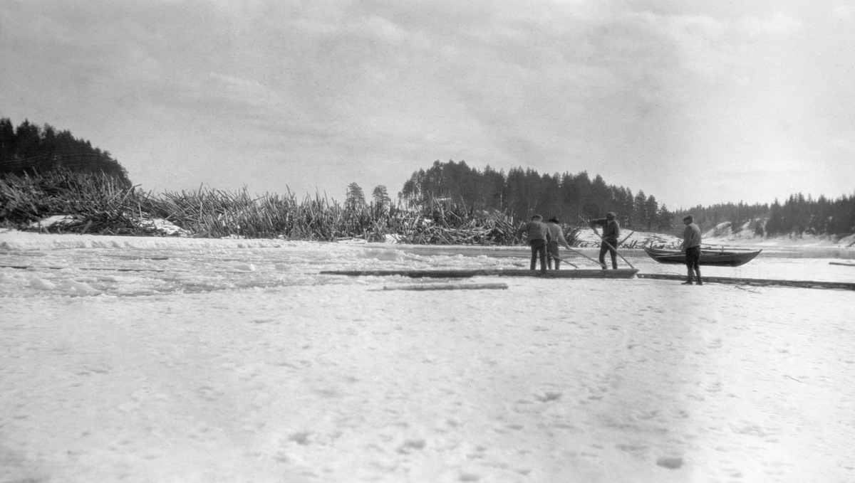 Fløterarbeid i Fossvika i Glomma, nedenfor Braskereidfossen i Våler kommune i Hedmark. Dette fotografiet ble tatt i april 1919, før isen hadde gått. I forgrunnen ser vi tre menn som trakk en tømmerstokk bortover isflata ved hjelp av fløterhakene sine, mens en fjerdemann sto og så på. Bak dem, på isflata mot den åpne råken under fossen, lå det en robåt. Karene forberedte antakelig sprenging av isen, slik at det skulle bli mulig å arbeide ut tømmeret i en haug som lå nederst i den bakenforliggende fossen før denne sesongens fløtingsvirke kom drivende med strømmen - jfr. SJF.1990-01272.
