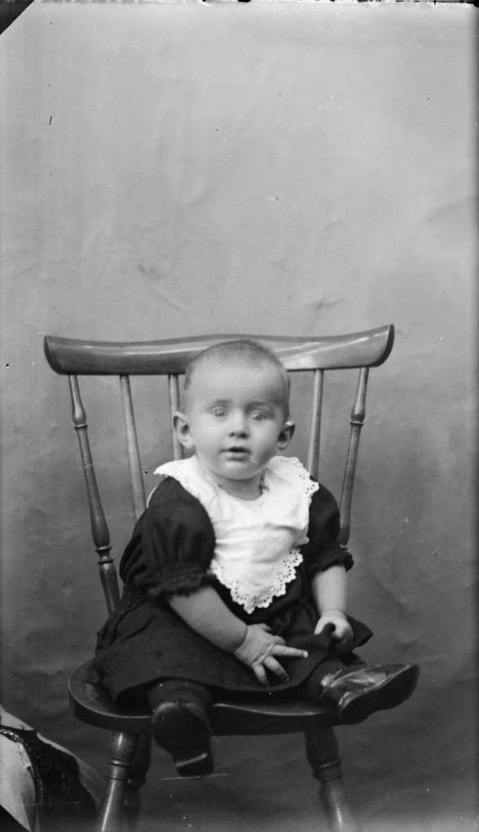Barn anttat 1år som sitter på en pinnestol. Hun har kort hår og mørk kjole med hvit krage