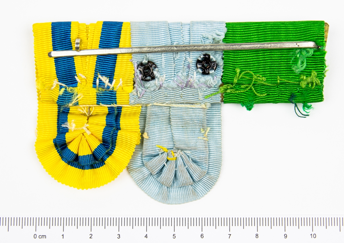 Medaljhållare i metall och tyg. Tygets färger grön, ljusblå samt blå/gul. Fäste på baksidan i metall.