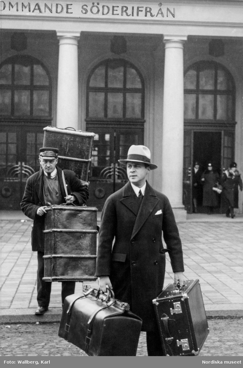Två män framför Stockholms central, båda bär på bagage, resväskor och koffertar. Den ene mannen är ett stadsbud, den andre Kurt Jacobsson, chef för NK:s franska damskrädderi.