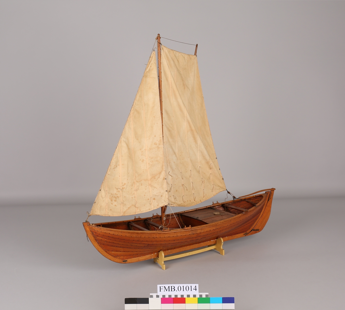 Modell av fiskebåt med brønn med  mast og 1 rombeformet seil og 1 trekantet. Tvers over båtens akterende er en stang hvor det rombeformete seilet er festet og som rorstangen kan hvile på.