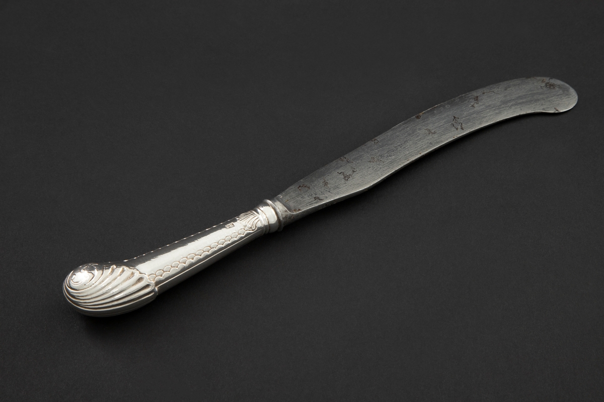 Kniv med sølvbelagt skaft og knivblad av stål. Knivbladet er flatt, buet med rund avslutning. Skaftet er rundt og dekorert med palmett- og bølgeornamentikk. Enden av skaftet er formet som et skjell med planteornamentikk.
