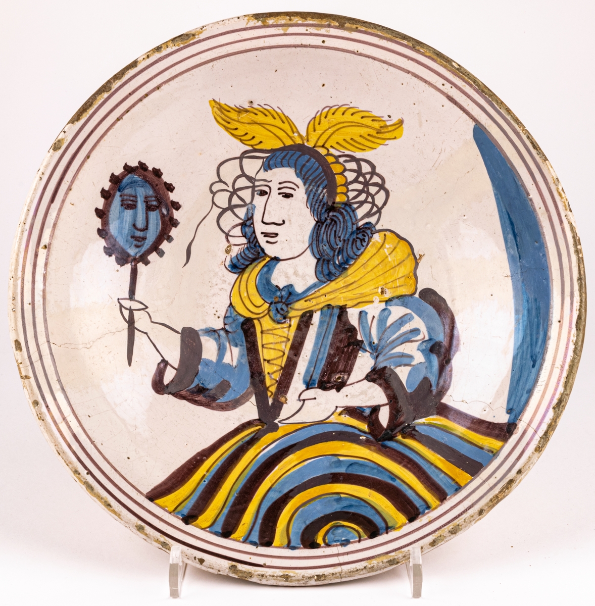 Keramikfat av ljusbrännande lergods med ljus tennglasyr som bottebfärg. Handdekorerad med kvinnogfigur som håller en spegel i högra handen. Kvinnofiguren har vingar på huvudet, snörliv och vid kjol. Figuren är i färgerna brun-lila, gult och blått.
Fatet är troligen från 1700-talet.
En spricka i fatet som löper genom en tredjedel, små naggar i kanten samt