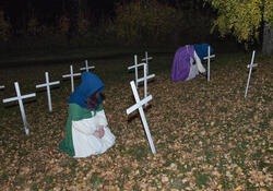 To koner med hetter langt fram over ansiktet sitter på en kirkegård og sørger, omgitt av hvite kors.