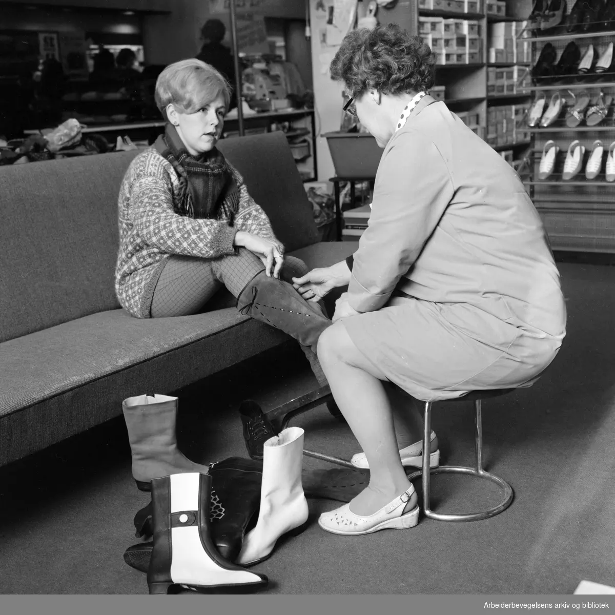 Skotøysalg. Berit Johansen (til venstre) prøver støvletter, assistert av Randi Thoresen i en skøtøyforretning i Storgata. Januar 1968