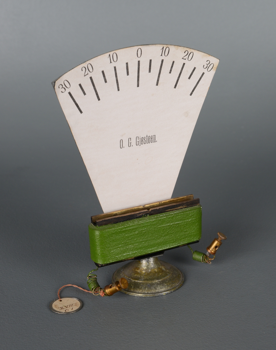 Et elektromagnetisk måleinstrument antakelig for å måle volt. En avlang spole med jernkjerne som står på en rund jernfot med hvit gradskive over. Instrumentet mangler målenålen. Gradeskalaen går fra 30 - 0 - 30. På gradskiven står det "O. G. Gjøsteen.".