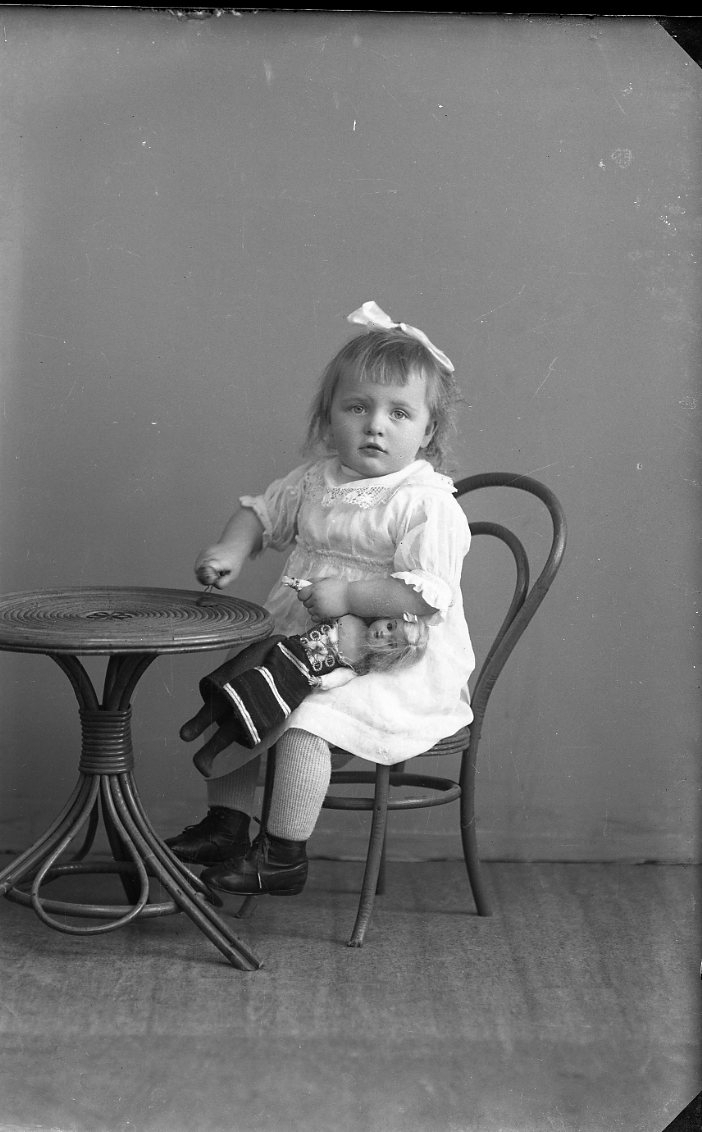En liten flicka med rosett i håret sitter i en barnmöbel med en docka i ena handen.