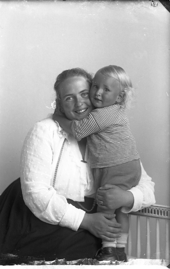 En leende kvinna sitter i en soffa med ett litet barn tätt intill sig. Barnet står på soffan och kramar kvinnan. Kvinnan är fru Johansson i Kumlaby på Visingsö.