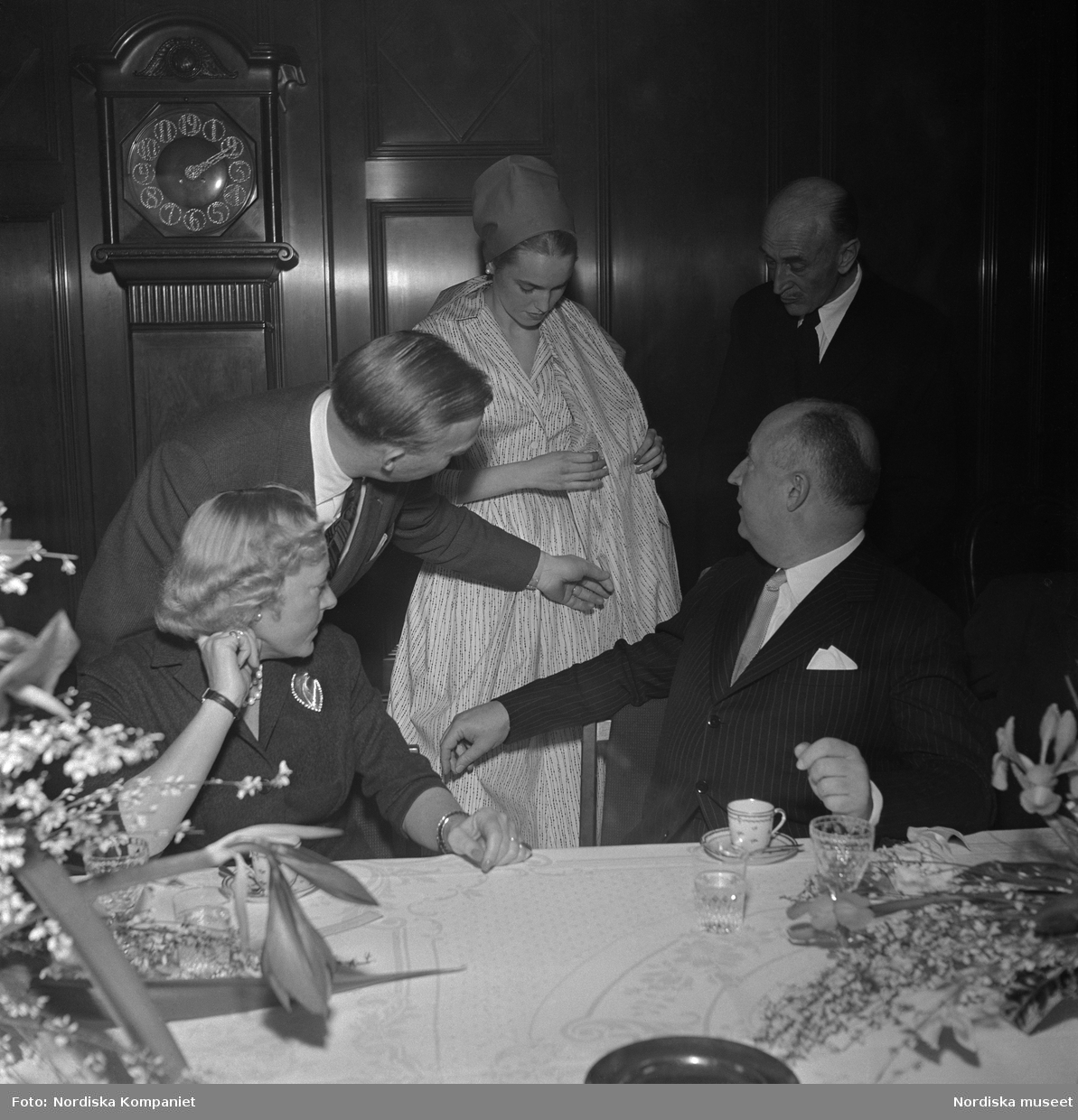 Christian Diors besök på NK, Nordiska kompaniet i Stockholm, februari 1957. Han bjuds på en visning av plagg uppsydda av tyger från NK:s textilkammare av Kurt Jacobsson och Pelle Lundgren. Mannekäng visar plagg. Vid bordet sitter Dior tillsammans med Astrid Sampe.
