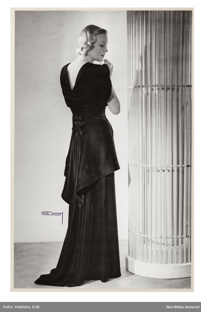 Mannekängen Margit Langlet poserar iklädd hellång, svart sammetsklänning med kort, draperad ärm och skört med snörning, sedd bakifrån. Från NK:s Franska damskrädderis egen ateljé. Klänningen framifrån se NMA.0099242.