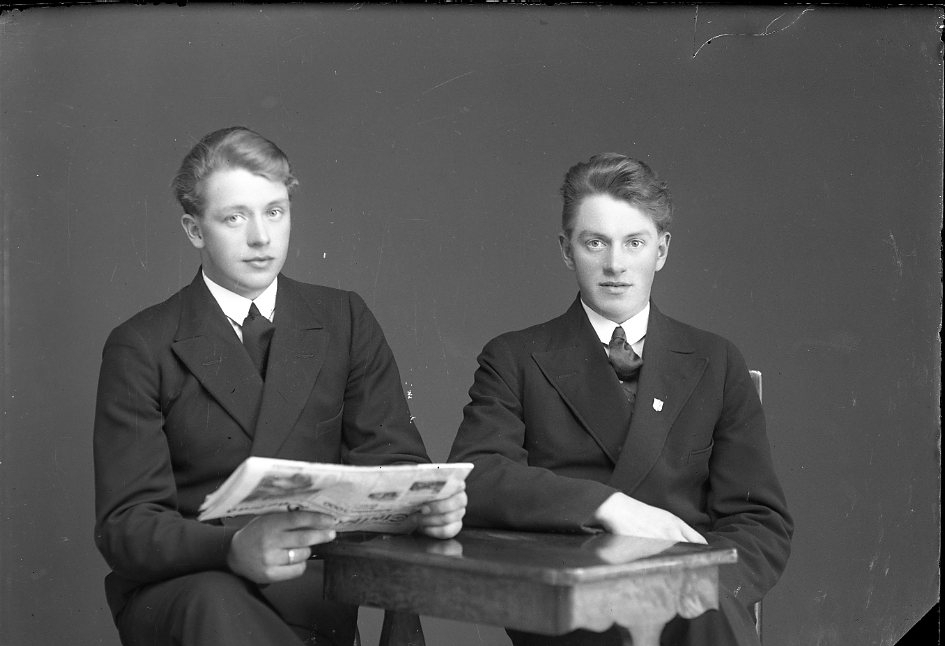 Gruppbild med två unga män som sitter vid ett bord, en av dem med en tidning i händerna. Mannen till höger är Evert Wibeck i Reaby.