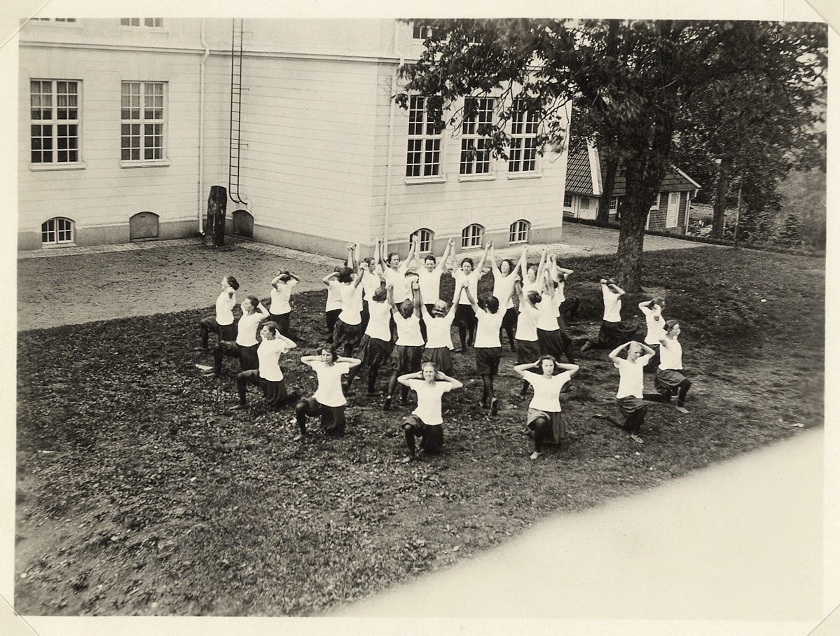 En grupp kvinnliga elever på folkskollärarseminariet (Ringsbergsskolan) i Växjö har gymnastik bakom skolan. Ca 1914.
I bakgrunden skymtar skolans vaktmästarbostad.