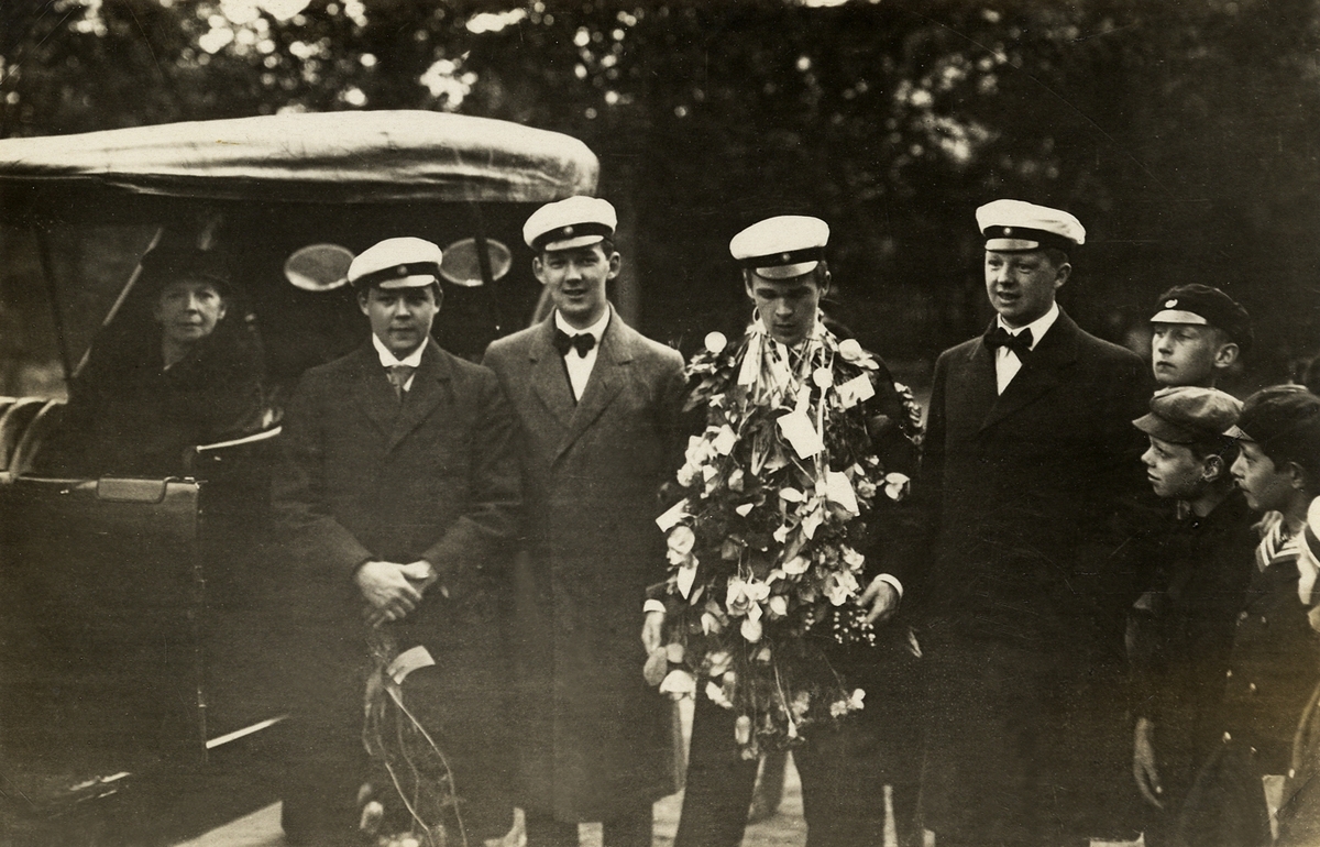 Tre ungherrar i rock och studentmössa uppvaktar en fjärde herre på hans studentdag. Till vänster sitter en kvinna i en bil och till höger beskådas det hela av fyra nyfikna pojkar. Ca 1920.