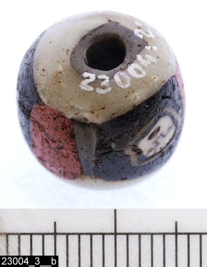 Anmärkningar: Kolbäck sn, Åsen 1:2 1:3 undersökt 1970.
Ansiktspärla, från gravfält daterat till äldre romersk järnålder.

Mosaikpärla av glas. Rund, något tillplattad mot hålen. Pärlan är uppbyggd kring en stomme av benvit glasfluss. Närmast runt hålet nedtill (ej upptill) en gråfärgad massa (bild 23004_3__b). Runt pärlan löper horisontellt ett 6-9 mm brett band indelat i tre svarta fält genom vertikala ränder i rött. I varje svart fält finns en ansiktsmask (bild 23004_3__c-e) i vitt av samma massa som stommen. Ansiktskonturerna markeras av en brungul slinga (möjligen hår) som är öppen nertill mot en tilltänkt hals. Ansiktena är något oregelbundna, men snarast av avrundat fyrkantig form. De är något snedställda åt olika håll och är av olika storlek, ca 3-5 mm. Olikheterna beror troligen på deformation i samband med tillverkningen. Ögonbryn, näsa och näsborre markeras av vinklade streck. Munnarna har oval form och är röda med svart kontur. Ögonen är markerade med svart kontur och har en markerad svart pupill på kritvit botten. Fynd 11 från anläggning A23 på gravfält RAÄ 191:1.
D:15 H:14

Pärlor av denna typ (Selling typ I) anses vara tillverkade på alexandrinskt eller syriskt område under första århundradet e.Kr. Om Kolbäckspärlan är tillverkad vid den tiden har den troligen inte nerlagts i graven före ca 100 e. Kr. De övriga pärlorna i grav A23 kan även de dateras till romersk järnålder.

Litteratur:
Magnusson, G. 1970. En mosaikpärla med ansiktsmasker
från Kolbäck. Fornvännen 1970, s. 227ff.
Samhör med osteologiskt material inom huvudnummer vlm-30419