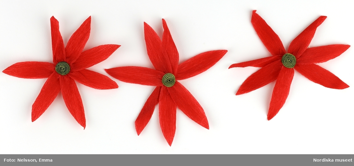 a-l) Tolv stycken hemtillverkade julgransprydnader av rött kräpp-papper i form av blommor/julstjärnor, med upphängningsanordning av knappnålar. 

Lena Kättström Höök 2019-03-21