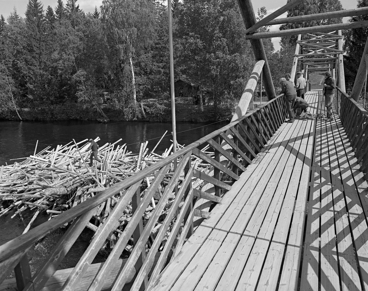 Tømmerhauger på motstrøms side av Klokkerfossbrua over Glomma i Elverum i Hedmark. Brua forbinder den østre elvebredden med Prestøya, der Norsk skogmuseum har sine friluftssamlinger. Klokkerfossbrua ble bygd i 1963-64 i en såkalt tømmersprengverkskonstruksjon med fire 20 meter lange spenn som kvilte på landkar i ytterendene og tre støpte betongkar i sjølve elveløpet. Under tømmerfløtinga var slike kar hindringer som hadde lett for å «fange» løstømmeret som kom drivende med strømmen. På denne måten kunne det bygge seg opp digre hauger eller tømmervaser på motstrøms side av brukarene, noe som hadde skjedd her. Egentlig skulle det ikke gå tømmer i Klokkerfossen i det hele tatt. Fløterne brukte å legge ei ledelense fra den østre elvebredden ved Elverum prestegard, noen hundre meter høyere oppe, mot et steinskjær i den sentrale delen av elveløpet. Derfra pleide «draget» fra Prestfossen på den nordvestre sida av Prestøya å være såpass kraftig at løstømmeret som kom fra ovenforliggende skogbygder strøk den vegen. Der var vannføringa større og faren for haugdannelser atskillig mindre. Ved denne anledningen må lensa ha sviktet, noe som førte til at fløterne fikk mye ekstraarbeid i Klokkerfossen. På dette bildet ser vi at en av karene fra fløterlaget hadde klatret ned på tømmerhaugen med et tau mens arbeidskameratene fortsatt befant seg på brua. Fotografiet ble tatt våren 1982.