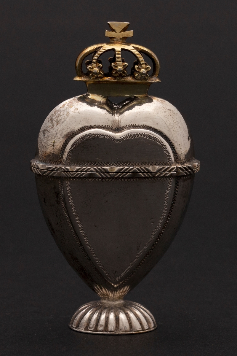 Hjerteformet luktevannshus i sølv med oval fot og forgylt krone på lokket. Hjerteformede felt på for- og bakside av korpus. Enkel dekor. Innvendig forgylt.